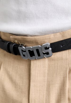 Ceinture tissée noire avec une grande boucle en métal argenté arborant les lettres "GCOS." Portée sur un pantalon en tissu beige.