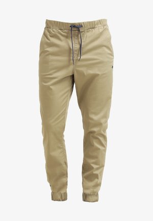 Hombre con camiseta gráfica blanca, pantalones jogger beige, zapatillas negras y mochila negra, caminando con una mano en el bolsillo.