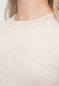 Gros plan d'une personne portant un haut côtelé beige clair avec un col rond, mettant l'accent sur le cou et la partie supérieure de la poitrine.