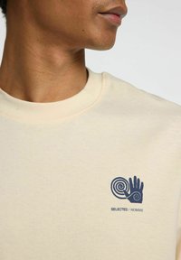 Beige bomullströja med rund halsringning, prydd med en marinblå spiral och handgrafik på vänster sida. Slät textur, avslappnad design.