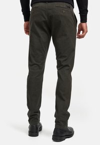 Pantalones de pana verde oscuro con corte recto, que presentan dos bolsillos traseros con botones y un acabado texturizado. Llevados con zapatos negros.