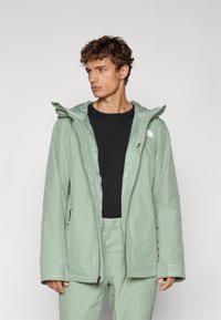 Chaqueta verde claro, impermeable, con frontal con cremallera y capucha ajustable. Presenta bolsillos laterales y puños texturizados. Forro interior negro.