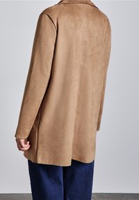 Persona vistiendo un abrigo largo de ante color beige y pantalones azul oscuro, visto desde atrás contra un fondo gris claro liso.
