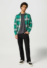 Cămașă din flanel cu carouri verzi și albe, purtată cu tricou negru, pantaloni negri și adidași negri. Cămașa are închidere cu nasturi și mâneci lungi.