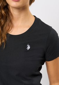 Černé tričko z měkké bavlny. Má kulatý výstřih a krátké rukávy, s malým bílým vyšívaným logem na hrudi.