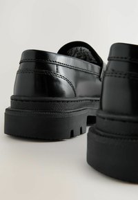 Czarne lakierowane buty na grubym, gumowym podłożu, z zaokrąglonym noskiem i widocznymi detalami przyszycia. Gładka faktura z eleganckim wzornictwem.