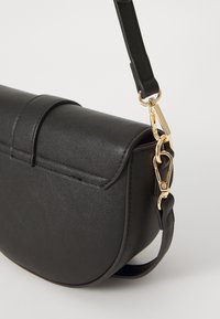 Bolso cruzado de imitación cuero negro con forma curva, herrajes en dorado, correa ajustable y detalle de cierre con solapa. Textura suave.