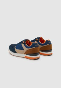 Par de zapatillas deportivas en azul, beige, marrón y naranja, mostradas en ángulo para destacar los detalles traseros y laterales sobre un fondo claro y liso.