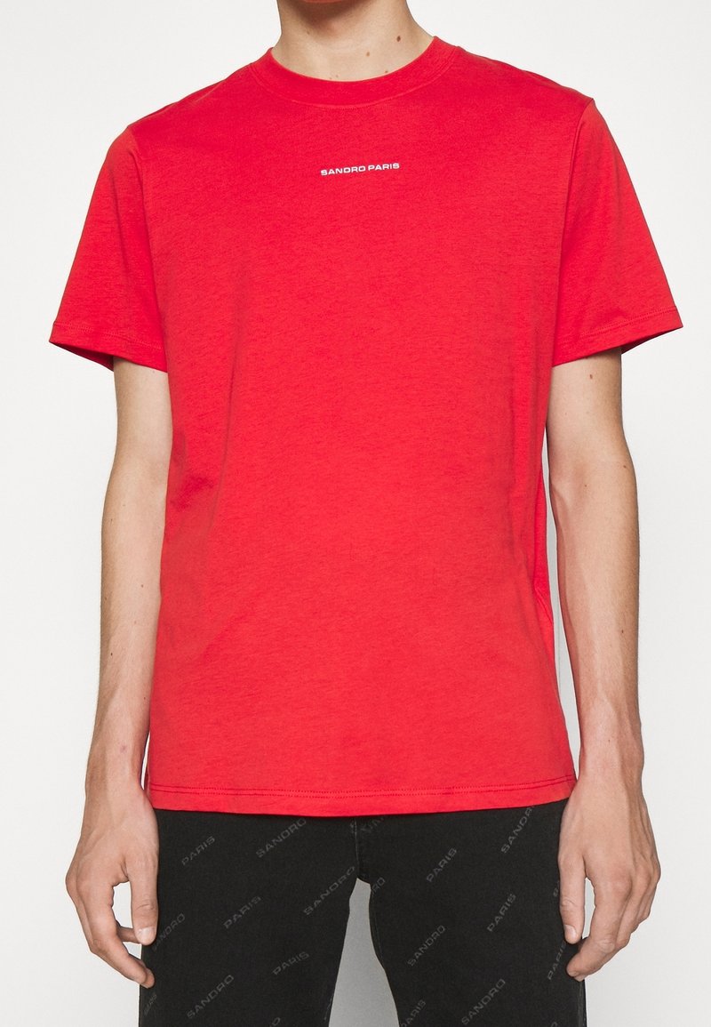 T-shirt rosso a maniche corte in cotone, con colletto rotondo e logo "Sandro Paris" in bianco sulla parte anteriore. Texture liscia.