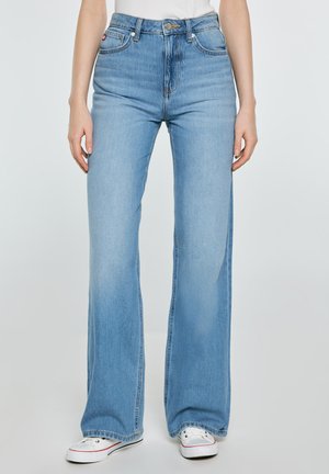 Kobieta nosząca jasnoniebieskie jeansy z wysokim stanem i szerokimi nogawkami, w połączeniu z białymi trampkami i białą bluzką, stojąca z rozluźnionymi ramionami wzdłuż ciała.