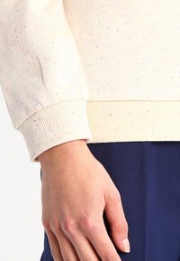 Sweatshirt de couleur crème avec des éclats colorés, finitions côtelées aux poignets et à l'ourlet. Le matériau semble doux, avec une finition texturée visible aux coutures.