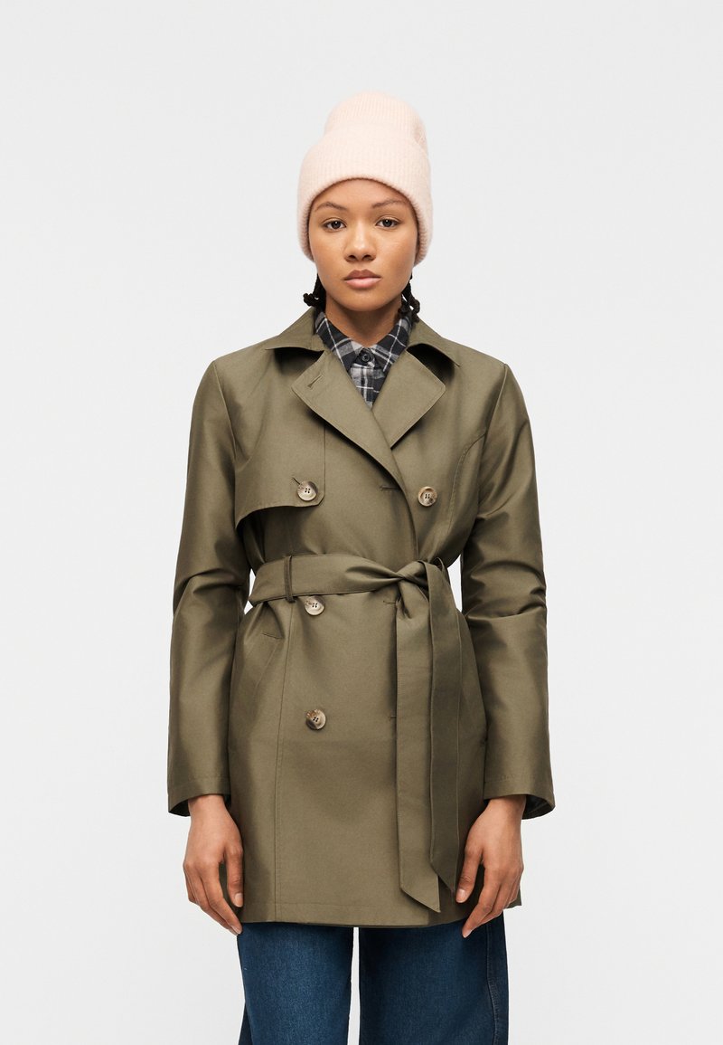 Γυναίκα που φοράει μπεζ πλεκτό καπέλο, χακί παλτό τύπου trench με ζώνη και μπλε τζιν, στέκεται μπροστά σε απλό φωτεινό φόντο.