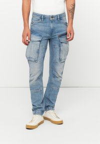 Man wearing light blue denim cargo jeans with multiple pockets and white sneakers against a plain white background.