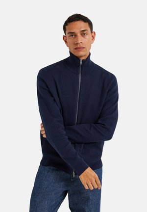 Pull zipé marine avec col montant et poignets côtelés, fabriqué à partir d'un matériau doux. Porté sur un jean en denim, il présente une texture lisse.