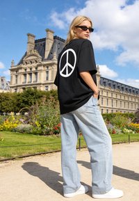 Jeune femme portant un t-shirt noir avec un symbole de paix, jean bleu clair et lunettes de soleil, debout dans un jardin près d'un bâtiment historique sous un ciel bleu.
