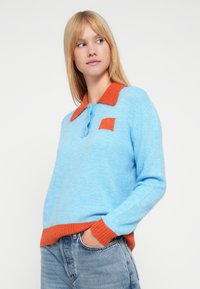 Hellblauer Pullover mit orangefarbenem Kragen und Manschetten, mit einem quadratischen Aufnäher und drei Knöpfen vorne, aus weichem Strickmaterial gefertigt.