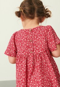 Rotes Blumenkleid mit kurzen Ärmeln, Knopfverschluss auf der Rückseite und gerafftem Taillenbereich. Es hat ein strukturiertes Gewebe mit weißen Blättermustern.