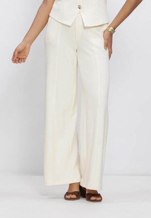 Mujer con pantalones blancos de talle alto y pernera ancha y sandalias marrones de tacón con los dedos al aire, con una mano en la cadera y una pulsera visible en la muñeca.