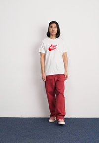 Balta marškinėliai su raudonu Nike logotipu, derinami su laisvai krentančiomis raudonomis kelnėmis. Modelis stovi ant mėlynos kiliminės dangos priešais baltą sieną.
