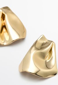 Bijoux ton doré au design fluide et sculptural, présentant des surfaces lisses et réfléchissantes ainsi qu'une forme légèrement ondulée.