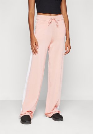 Vrouw met lichtroze wijde broek met witte zijkanten en zwarte instapschoenen, staand tegen een effen witte achtergrond.
