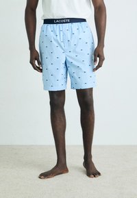 Light blue shorts featuring a patterned design with small dark blue crocodiles. Elastic waistband with "LACOSTE" branding. Smooth texture.