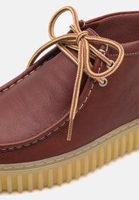 Brown leather shoe with a textured surface, featuring yellow and brown laces, and a chunky, ridged beige sole.