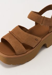 Pruuni nahast sandaalid, millel on kaks laia rihma, jäme platvormsolg ja tekstureeritud viimistlus. Kerge UGG bränding külgedel.