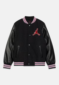 Černá varsity bunda s černými koženými rukávy, červenými a bílými pruhovanými manžetami a červeným vyšívaným logem Jumpman na hrudi. Obsahuje patentky a boční kapsy.
