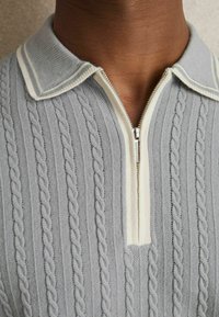 Pulover tricotat gri deschis cu guler cu fermoar, având accente crem, textură ribată și modele decorative din cabluri.