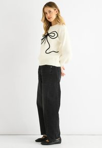 Vit stickad tröja med en svart rosett-design, i kombination med högmidjade svarta jeans och svarta ballerina-skor. Slät textur, avslappnad passform.