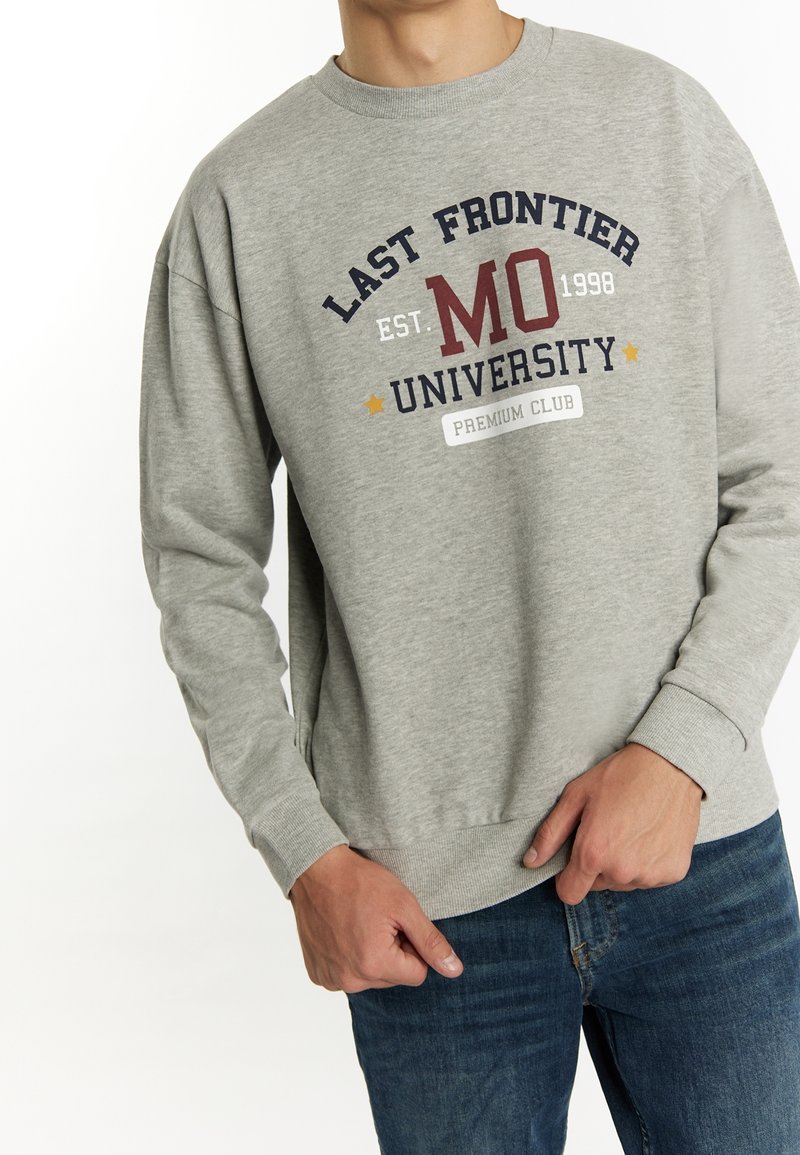 Homme portant un sweat gris avec le texte "Last Frontier MO University Est. 1998 Premium Club" et un jean bleu, se tenant avec les mains près de la taille.
