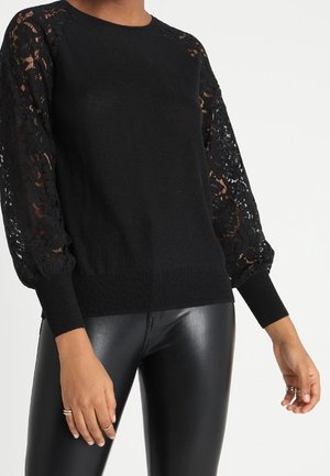 Pull en maille noir avec manches ballon en dentelle florale, associé à un pantalon en cuir noir sur une personne debout.