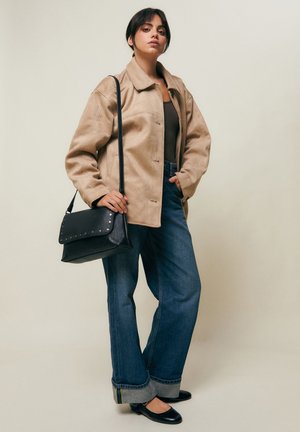 Noor naine lühikeste tumedate juustega, kandes beeži jopet, tumedat topi, ülesrullitud siniseid külgi, musti baleriinikingi ja musta õlakotti, tavalise tausta ees.