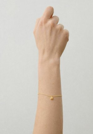 Kohotettu käsivarsi, jossa on herkkä kultainen ketjukoru pienen pyöreän riipuksen kanssa, yksinkertaista vaaleanharmaata taustaa vasten.