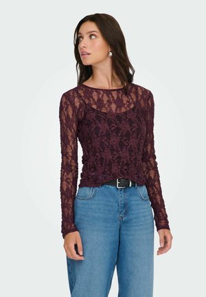 Burgunderfarvet blonde top med lange ærmer, der har et blomsterdesign og en rund halsudskæring, parret med højtaljede blå jeans og et sort bælte.