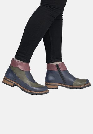Person trägt schwarze Hose und bunte Stiefeletten mit blauen, olivgrünen und pinkfarbenen Lederabschnitten, steht mit überkreuzten Füßen.