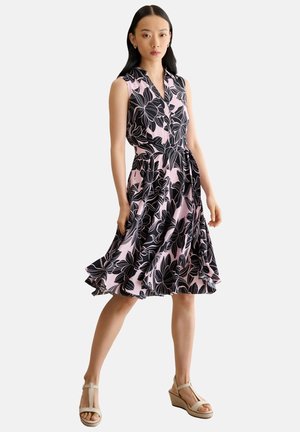 Mujer con un vestido rosa sin mangas con un gran estampado floral negro, cinturón en la cintura y sandalias beige con cuña, de pie frente a un fondo blanco.