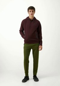 Sudadera marrón con cordón, bolsillos laterales; combinada con pantalones oliva ajustados. El modelo se encuentra en posición neutra, llevando zapatos negros.