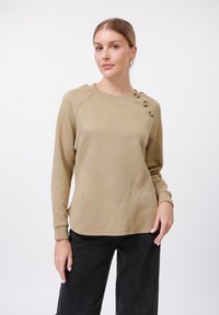 Felpa beige a trama con motivo geometrico, scollo rotondo e tre bottoni decorativi su una spalla. Abbinata a pantaloni neri a gamba larga.