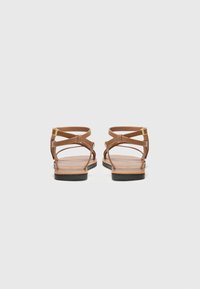 Bruine leren sandalen met kruisende banden, platte zool en kleine gouden hardware-accent. Minimalistisch ontwerp met open achterkant en tenen.