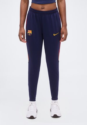 Personne portant un pantalon de sport FC Barcelone bleu marine avec logo Nike jaune et bandes latérales rouges, associé à des baskets blanches.