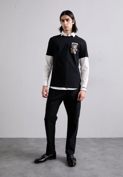 Black t-shirt with a teddy bear print and "Moschino" text, layered over a white collared shirt. Black trousers and shiny black shoes complete the look.