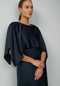 Námornícky hodvábny šaty s prehnutým overlay a širokými rukávmi, s lodičkovým výstrihom a hladkou textúrou. Modelka s jemne prekríženými rukami.