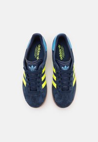 Modré semišové tenisky se třemi fluorescenčně žlutými pruhy, gumovou podrážkou a světle modrými detaily. Vybavené polstrovaným límcem a texturovanou stélkou.