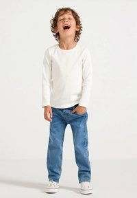 Krullend kind lacht met gesloten ogen, draagt een wit shirt met lange mouwen, blauwe spijkerbroek en witte sneakers, staat met één hand in de zak.