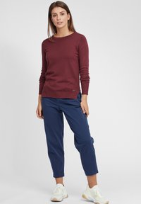 Maglione lungo bordeaux a maniche lunghe con scollatura rotonda, polsini aderenti e dettagli di cucitura laterali, abbinato a pantaloni tapered blu navy e sneakers bianche.