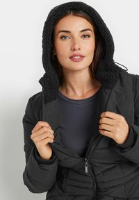 Giacca a vento nera con cappuccio foderato in pile dal design texture. Presenta una chiusura frontale con zip e una vestibilità aderente, indossata sopra un top scuro.