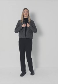 Szara kurtka na zamek z dwoma przednimi kieszeniami, noszona na czarnej bluzce i czarnych spodniach. Tkanina wydaje się mieć fakturę i być nieco gruba.