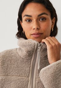 Veste en polaire beige floue avec un col montant et une fermeture éclair. Présente des accents de tissu lisse et léger le long de la fermeture éclair et des poignets.